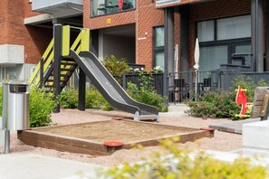 A playground with a slide and a small sandbox, surrounded by a brick building and a metal fence.