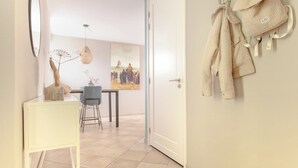 Un couloir avec une porte blanche, une table blanche avec des chaises, un porte-manteau suspendu et un tableau accroché au mur.