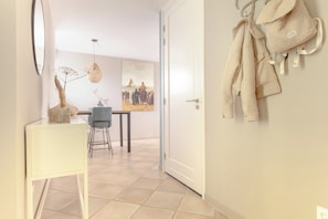 Een gang met een witte deur, een witte tafel met stoelen, een hangende kapstok en een schilderij aan de muur.