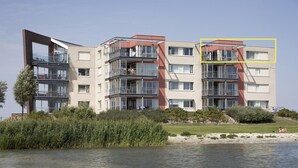 Un immeuble résidentiel à plusieurs étages, doté de balcons, d’un toit plat et offrant une vue sur une étendue d’eau.
