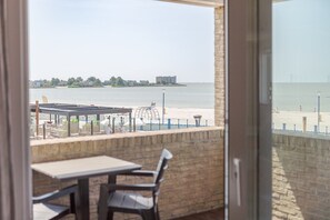 A balcony with a view of a beach and water, a table, and chairs.