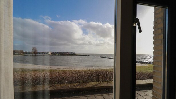 A view from a window overlooking a body of water, a grassy area, and a distant shoreline with buildings. - Lakeview Suite with Whirlpool in Makkum (Makkum)