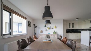 A dining area with a long table, chairs, a hanging light fixture, a window, and a kitchen in the background. - Lakeview Suite with Whirlpool in Makkum (Makkum)