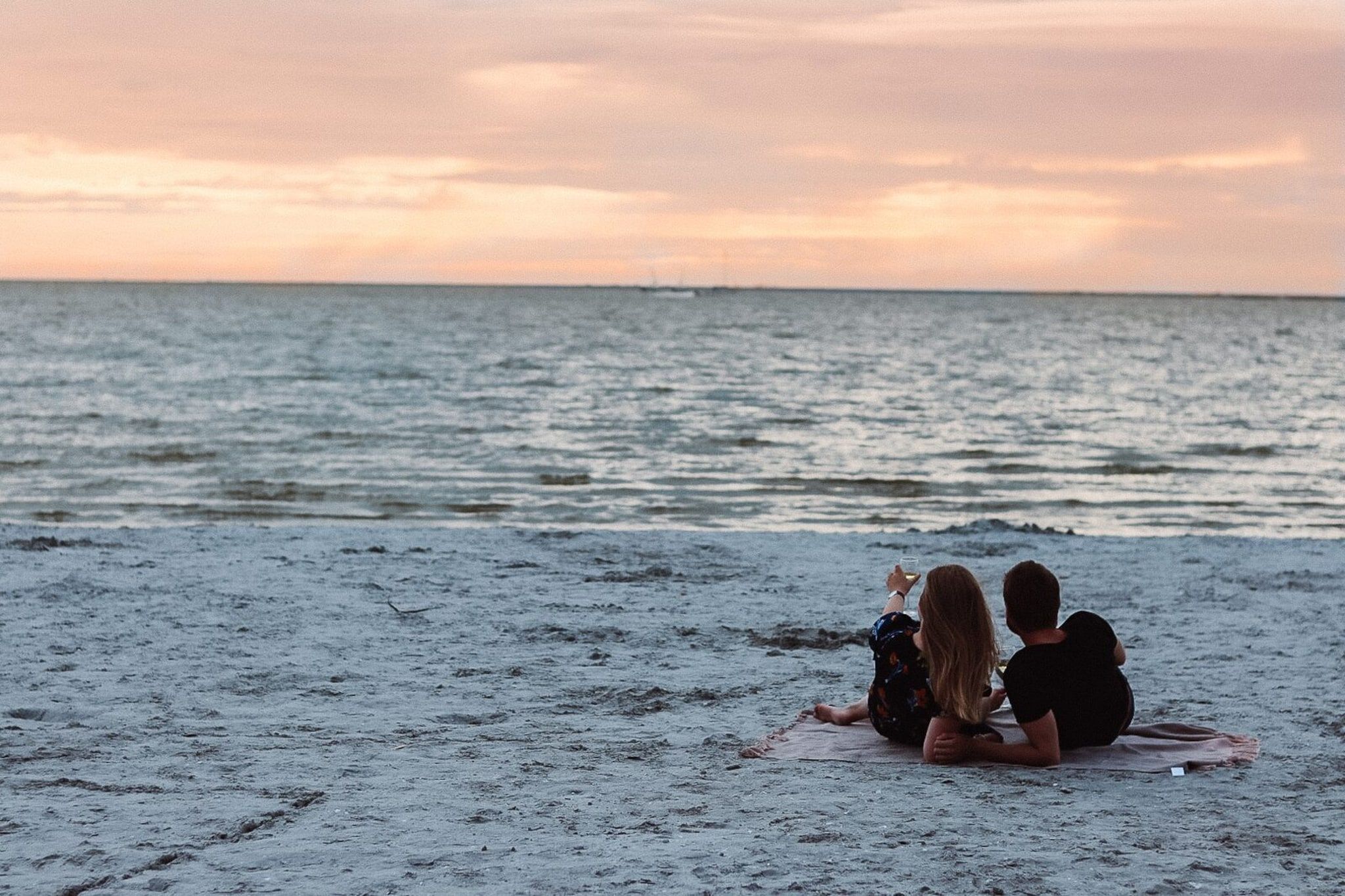 Two individuals sitting on a beach facing the ocean during sunset.