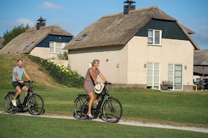 Two people riding bicycles on a path with thatched-roof buildings in the background.