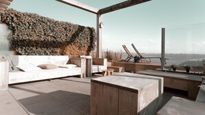 Un área de estar exterior moderna con vista al agua, que incluye un banco de madera, una zona de estar acolchada y una pared decorativa.