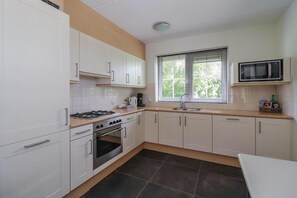 A compact kitchen with white cabinets, a gas stove, an oven, a microwave, and a window with blinds. - Waterfront Beach Villa for 5 (Makkum)