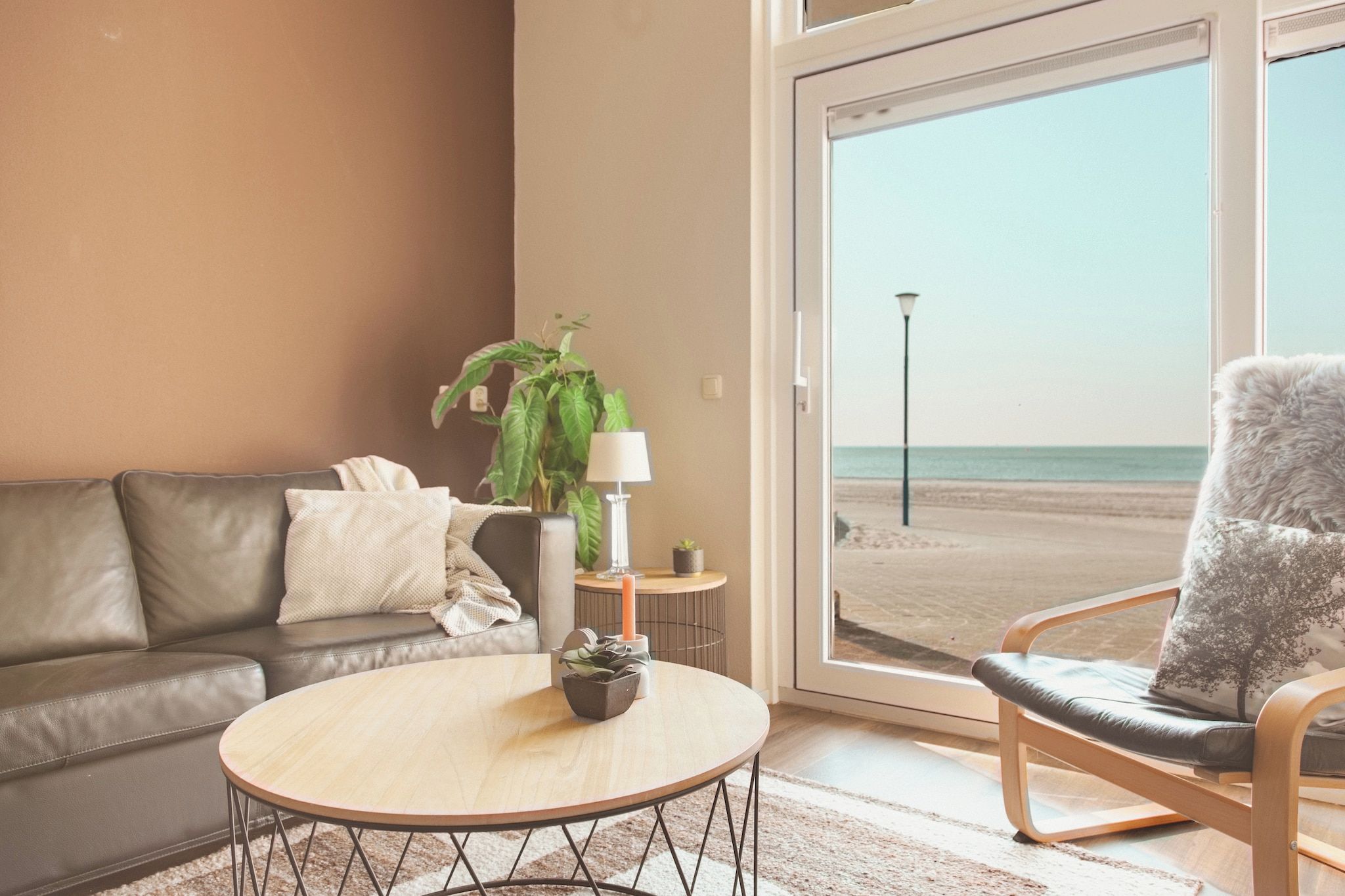 Een woonkamer met een leren bank, een rond houten bijzettafeltje, een potplant, een lamp en uitzicht op het strand door een groot raam.