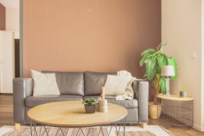 Een woonkamer met een grijze bank, een rond houten bijzettafeltje, een potplant en een lamp op een nachtkastje.