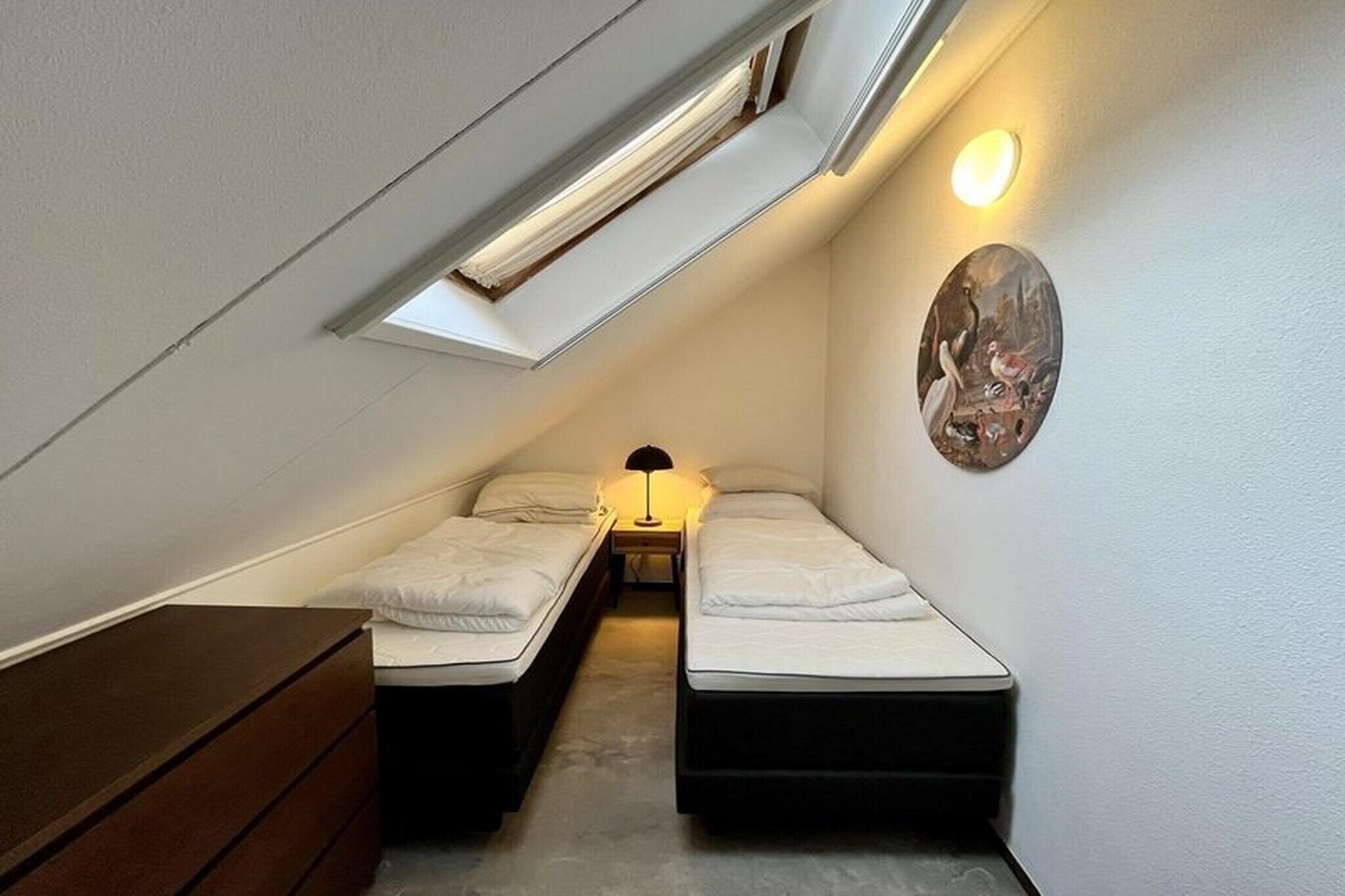 A small, narrow room with two beds, a nightstand, a lamp, and a wall-mounted artwork.