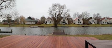 A waterfront property with a wooden deck, a dock, and a row of houses across the water.