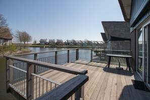 A wooden deck with a railing, a table, and a view of houses across a body of water. - Luxury Waterfront Wellness Villa (Makkum)