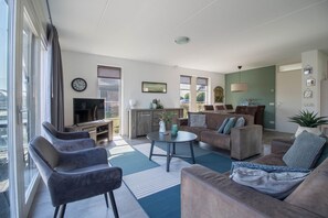 A modern living room with a sofa, chairs, a TV, and a kitchen area. - Luxury Waterfront Wellness Villa (Makkum)