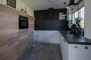A modern kitchen with wooden cabinets, a built-in oven, a black range hood, white drawers, and a window with a view. - Luxury Waterfront Wellness Villa (Makkum)