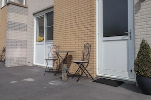 Ein Gebäudeeck mit einer weißen Tür, einem Fenster mit Insektenschutzgitter und zwei Metalltischen mit Stühlen.