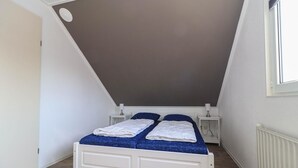 Une petite chambre blanche avec deux lits, un plafond en pente, une fenêtre et un radiateur.
