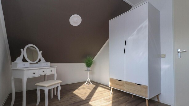 Une pièce avec une coiffeuse blanche, un petit tabouret, une plante en pot et une armoire agrémentée d’un élément décoratif en bois.