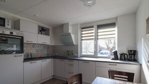 Une cuisine avec des meubles blancs, un pare-feu en carrelage, un four encastré, une fenêtre avec des stores et un coin repas avec une table et des chaises.