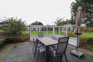 Eine Terrasse im Freien mit Tisch und Stühlen, einem Sonnenschirm und Blick auf ein Haus durch eine Glaskonstruktion.