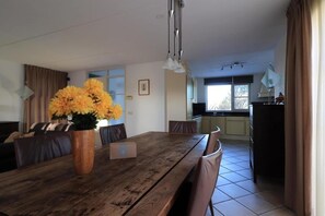 Ein Essbereich mit einem Holztisch, Stühlen und einer Vase mit gelben Blumen. Im Hintergrund ist eine Küche mit Schränken und einem Fenster zu sehen.