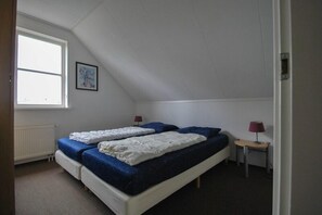 Een kamer met een schuine kap, twee bedden met blauw beddengoed, een klein tafeltje met een lamp en een radiator.