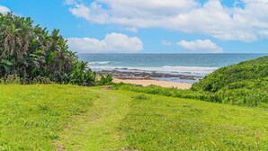 Paisagem costeira com primeiro plano gramado, praia de areia e oceano ao fundo.