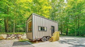 Une petite cabane à un seul étage, avec une entrée à l’avant et une porte sur le côté, située dans une zone boisée. Une allée de gravier mène à une rampe en bois qui donne accès à la porte.