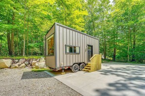 A small, single-story cabin with a front entrance and a side door, situated in a wooded area with a gravel driveway and a wooden ramp leading up to the door.