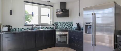 A modern kitchen with dark cabinetry, a stainless steel refrigerator, a built-in oven, and a window providing natural light.