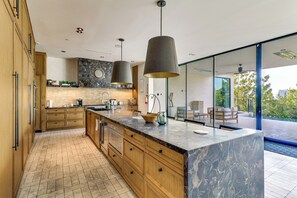 Eine moderne Küche mit Holzschränken, einer Marmorinsel, Pendelleuchten und Blick ins Freie.