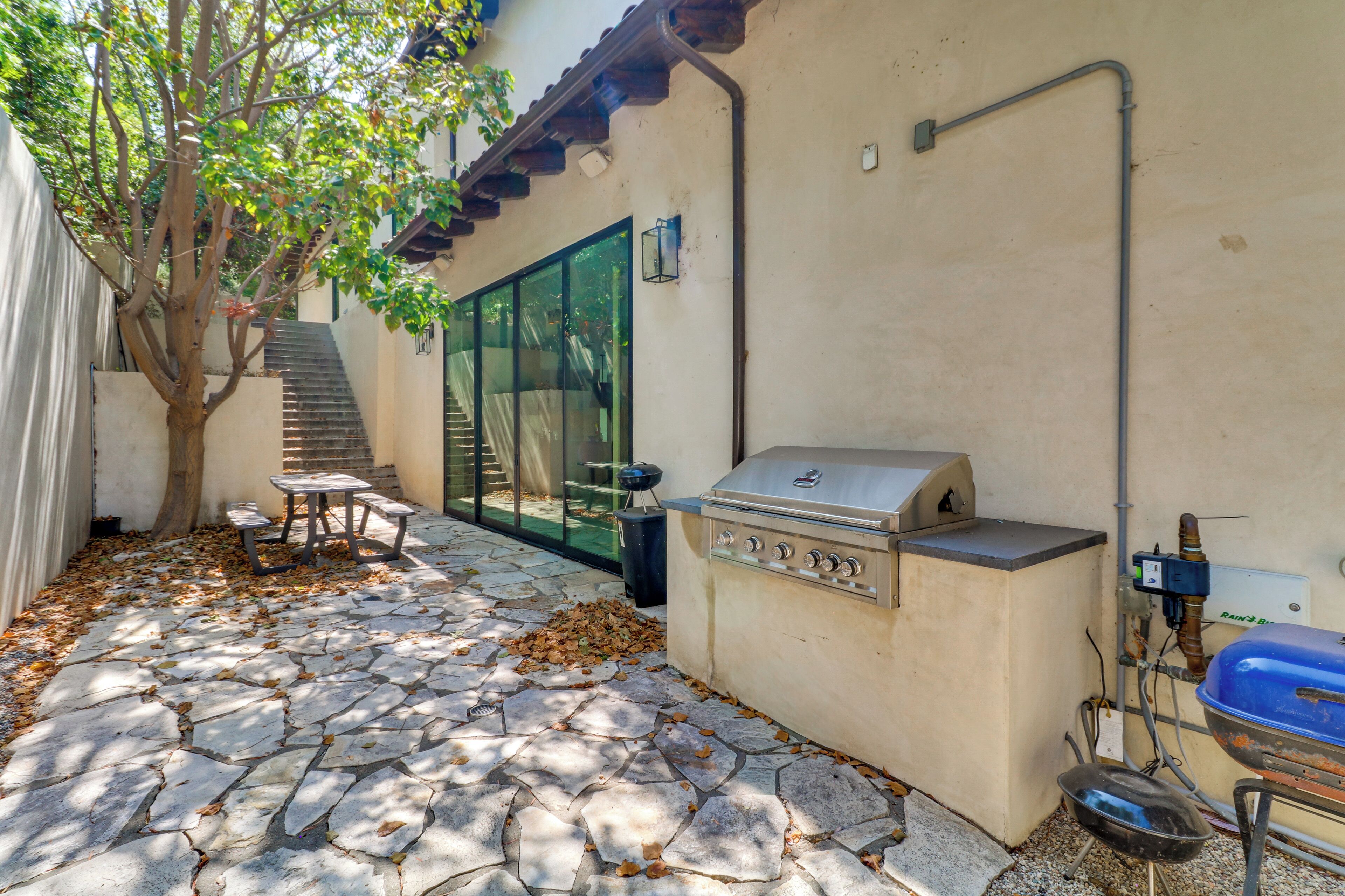 An outdoor patio with stone flooring, a barbecue grill, a table with chairs, and a tree.