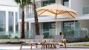 Un espace aménagé au bord de la piscine, avec des chaises longues et un parasol, des palmiers et un bâtiment moderne doté de grandes fenêtres.