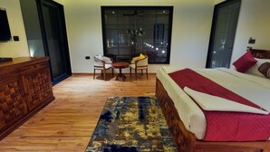 A bedroom with a bed, a desk, and a chair. There is a large window and a door. - Ranthambore Bunglow (Sawai Madhopur)