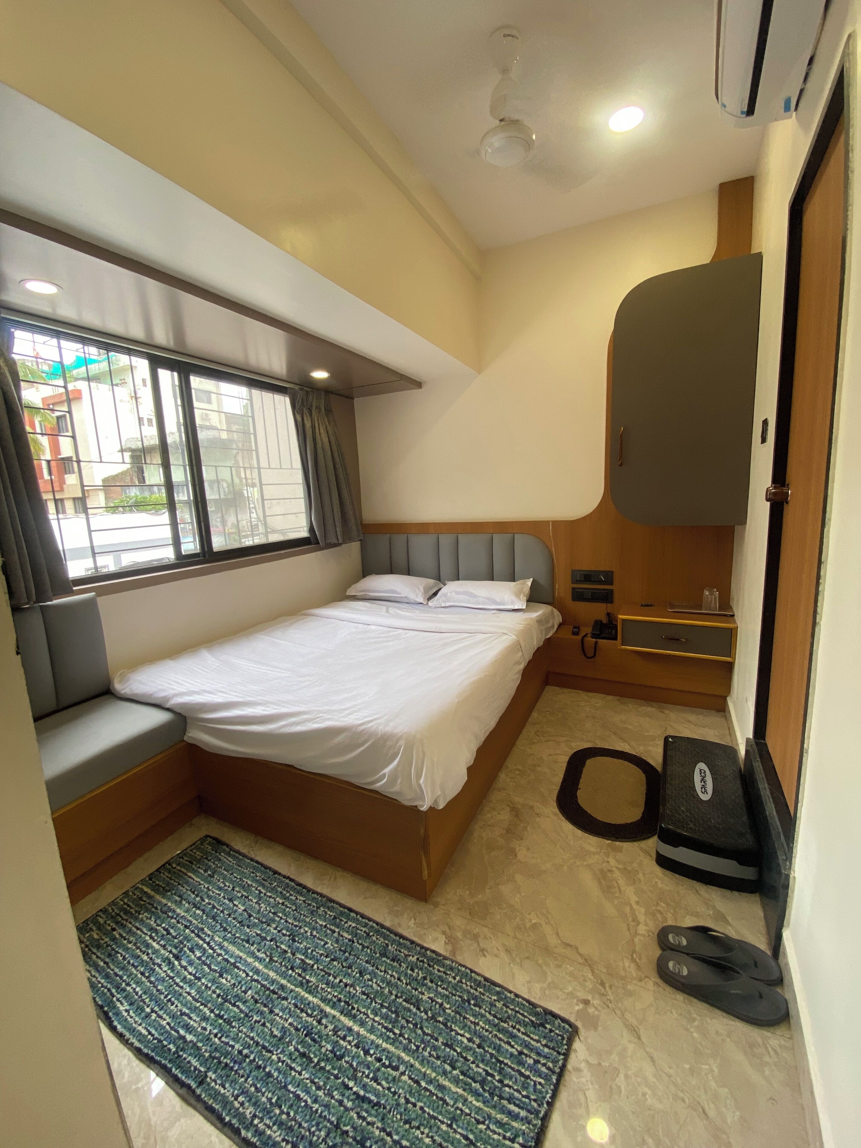 Una camera d'hotel con un letto, una finestra con tende, una scrivania con una sedia, un piccolo tappeto e una porta.