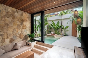 A modern interior with a stone wall, a wooden ceiling, a sofa, a TV, and a pool area visible through glass doors. - 1 BR Beachside Retreat Blacksand Villas Bingin (Badung)