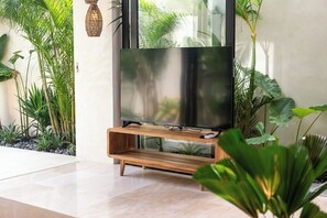 A modern living room with a flat-screen TV on a wooden stand, surrounded by lush green plants. - 1 BR Beachside Retreat Blacksand Villas Bingin (Badung)