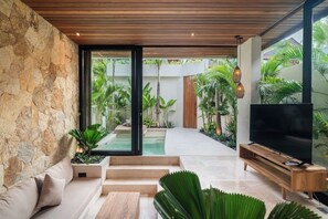 A modern interior with a stone wall, a wooden ceiling, a TV, and a seating area with steps leading to a pool. - 1 BR Beachside Retreat Blacksand Villas Bingin (Badung)