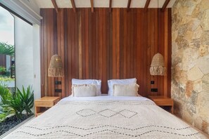 A bedroom with a wooden slat headboard, a bed with white bedding, two bedside lamps, and a view of greenery outside. - 1 BR Beachside Retreat Blacksand Villas Bingin (Badung)