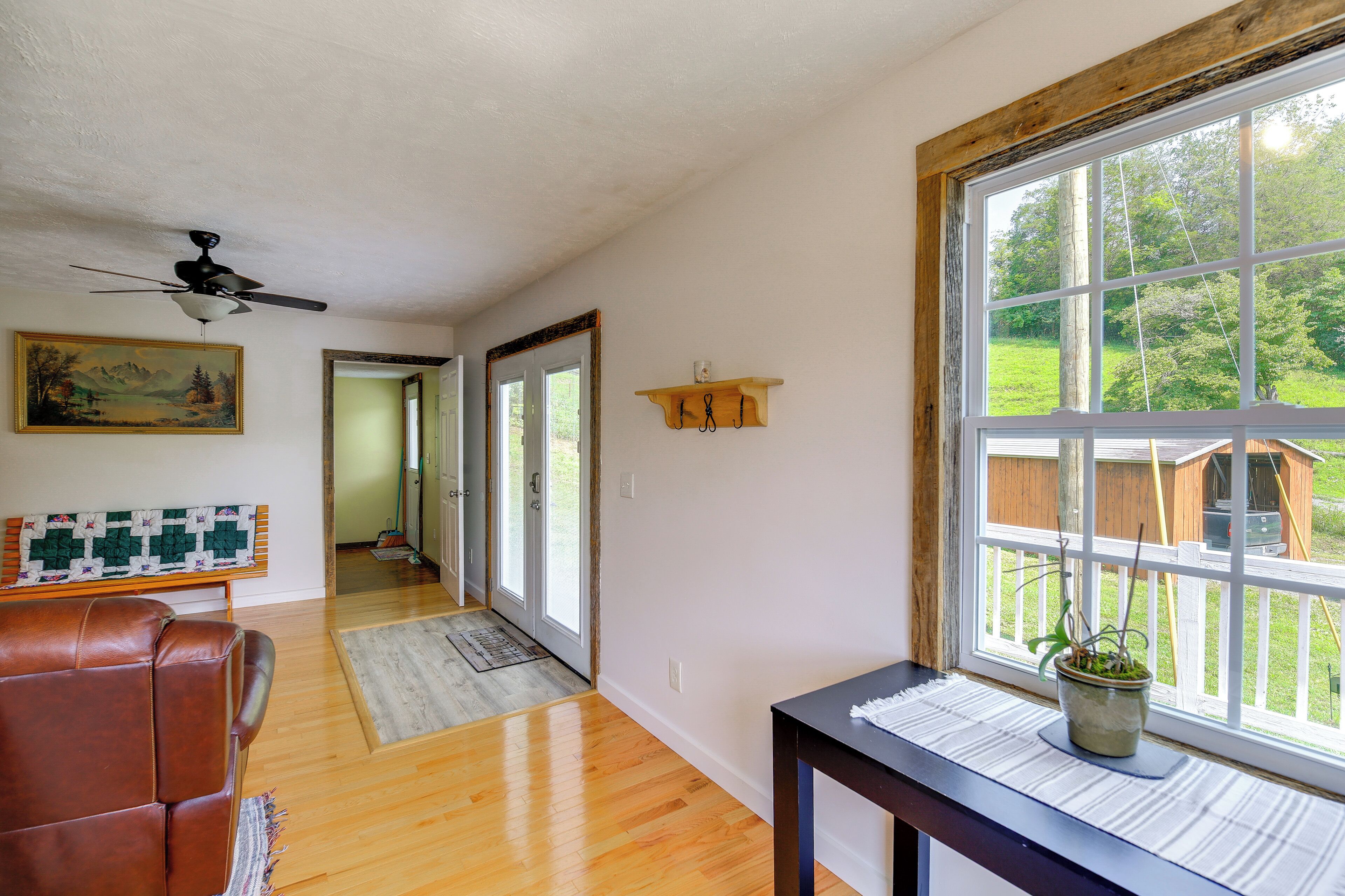 Una habitación con ventilador de techo, piso de madera, un sillón de cuero marrón, ventana con vista a un patio trasero y un cuadro en la pared.
