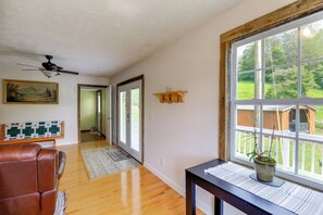 Une pièce avec un ventilateur de plafond, un plancher en bois, un fauteuil en cuir marron, une fenêtre donnant sur un jardin, et un tableau accroché au mur.
