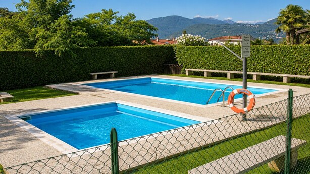Ein rechteckiger Pool mit Rettungsboje und Hinweisschild, umgeben von einem Zaun und Grünpflanzen.