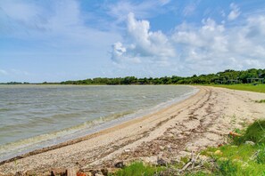 Ein Strand mit sandigem Ufer, ruhiger See und einer Baumreihe im Hintergrund.