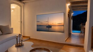 A modern living room with a large painting of a sunset over water, a white sofa, a coffee table with candles, and a view of a pool area. - Porto Cervo - Villa AUREA Retreat (Arzachena)