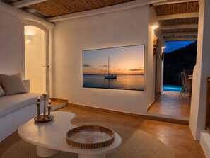 Ein modernes Wohnzimmer mit einem großen Gemälde, das einen Sonnenuntergang über Wasser zeigt, einem weißen Sofa, einem Couchtisch mit Kerzen und Blick auf einen Poolbereich.
