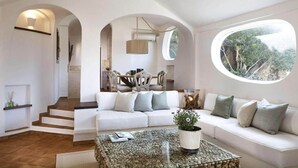 A modern living room with a white sofa, a coffee table with a plant, and a view through a large window. - Porto Cervo - Villa AUREA Retreat (Arzachena)