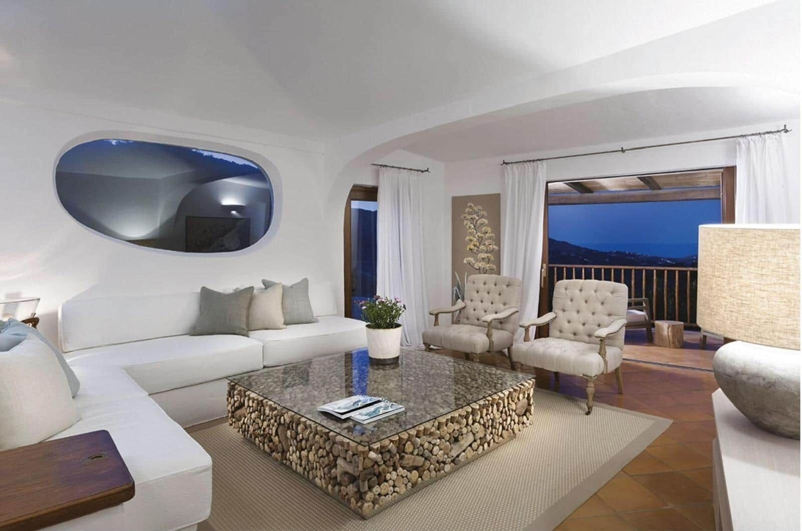 A modern living room with a white sofa, a glass coffee table with wood logs, and two armchairs.