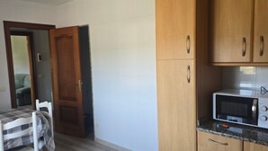 Een kleine keuken met een magnetron, houten kastjes, een eettafel met geruit tafelkleed en een deur naar een andere kamer.