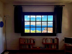 A room with a large window overlooking a coastal landscape, two bookshelves with books, a floor lamp, and a television. - Punta Est - PERLA Two-Bedroom Apartment with Terrace and Ocean View (Capo Coda Cavallo)