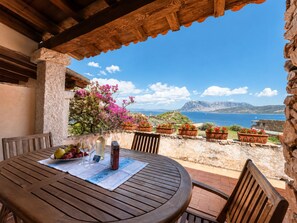 A wooden table with a placemat, surrounded by chairs, overlooking a scenic coastal view with potted plants and a clear sky. - Punta Est - PERLA Two-Bedroom Apartment with Terrace and Ocean View (Capo Coda Cavallo)
