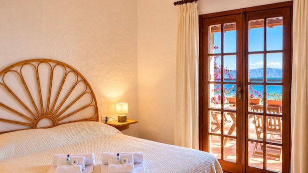 A hotel room with a bed, a nightstand with a lamp, and a view of the sea through a glass door. - Punta Est - PERLA Two-Bedroom Apartment with Terrace and Ocean View (Capo Coda Cavallo)